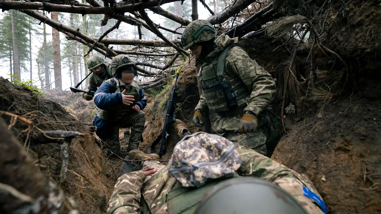 Drie Oekraïense militairen in een loopgraaf. Een tolk geeft instructies door bij een casus Zelfhulp Kameradenhulp (ZHKH). Een van de militairen neemt hierbij de rol aan van gewonde.