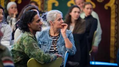 Twee mensen zitten naast elkaar in een zaal: een vrouw met grijs haar en een denim jasje luistert aandachtig, een man met lang haar kijkt mee.
