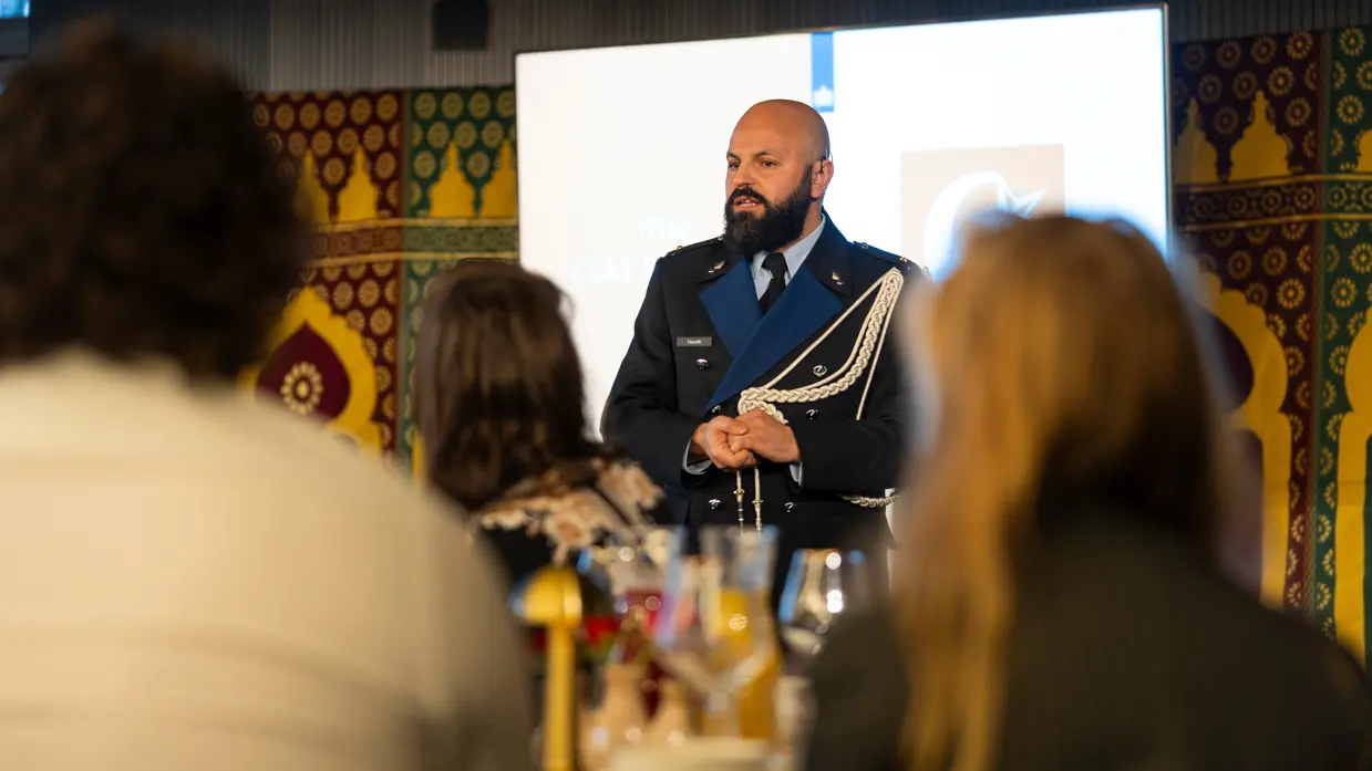 Links: Een man in een donkerblauw uniform met gouden versieringen staat voor een groep mensen en spreekt, terwijl het publiek naar hem luistert. Rechts een man met gesloten ogen die zingt in een microfoon. Hij houdt een handtrommel vast en draagt een donkerblauwe kleding, omringd door kleurrijke patronen.
