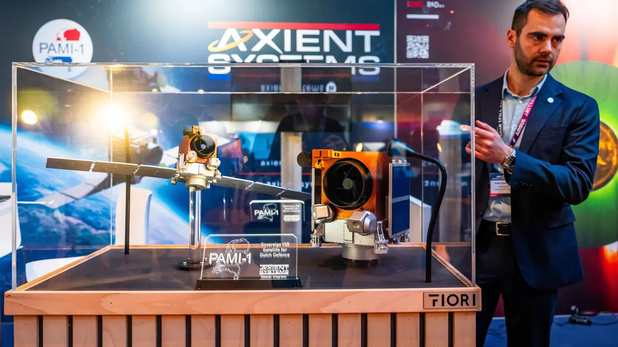 Links staat een man achter een vitrine waarin een schaalmodel is te zien van een satelliet. Op de andere foto is een schaalmodel van een andere satelliet te zien met gouden bekleding.