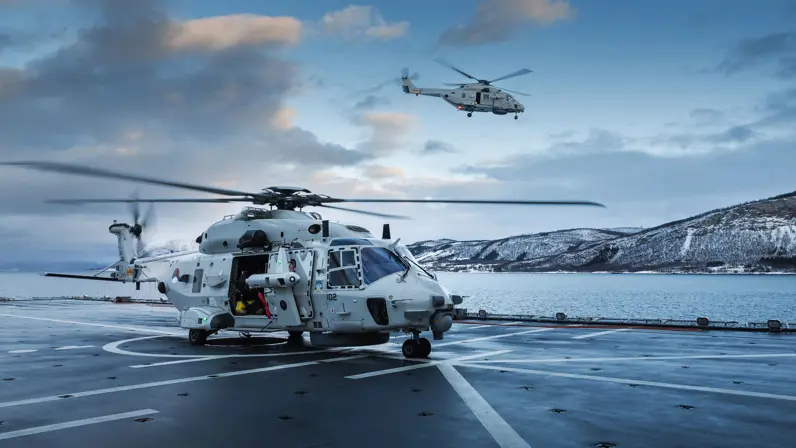 NH90-maritieme helikopter op het dek van een marineschip tijdens een oefening in een besneeuwd kustgebied, terwijl een tweede helikopter boven zee vliegt. 