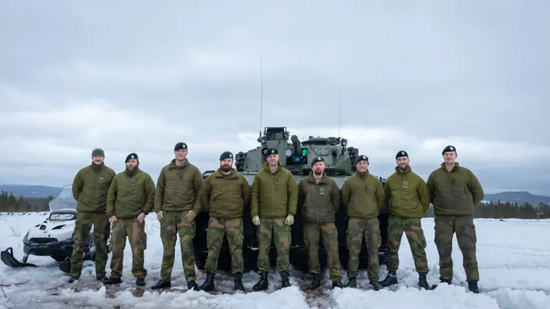 Het Noorse team van instructeurs opgelijnd in de sneeuw, voor een CV90 en een sneeuwscooter. 