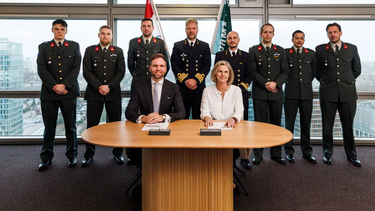 : Derk Boswijk naast een dame aan een ondertekeningstafel. Achter hen staan acht militairen van landmacht en marine. Ze staan voor de Nederlandse vlag en die van ABN AMRO.