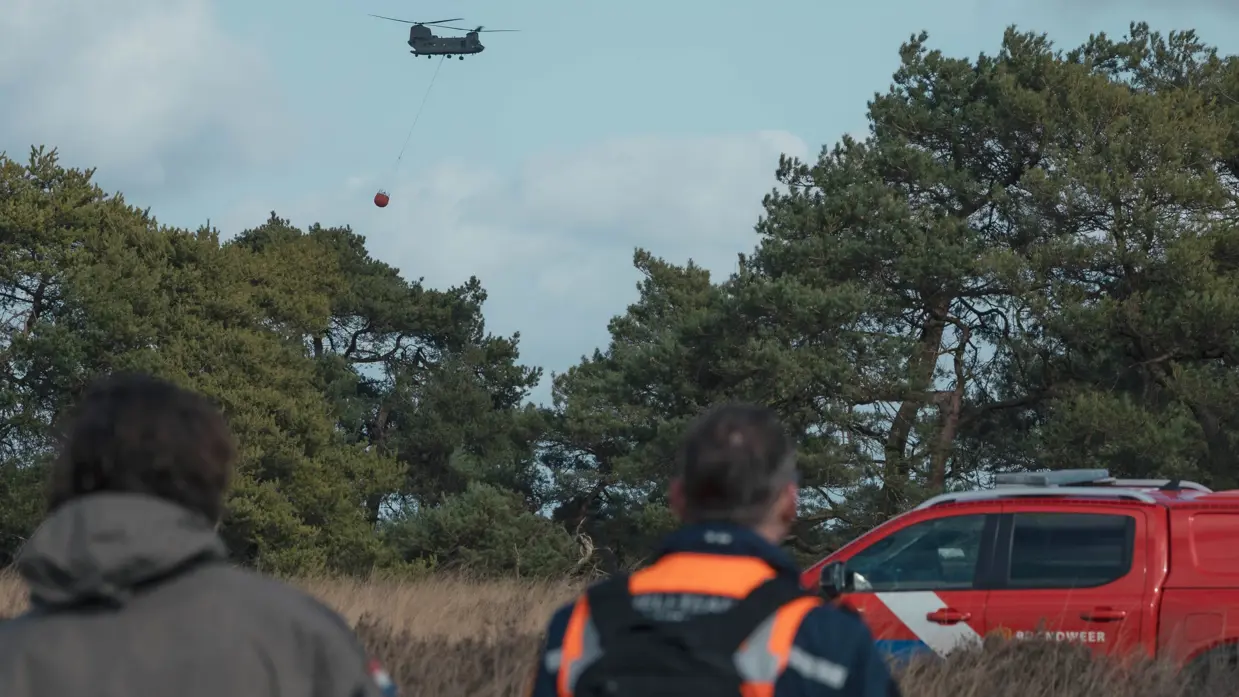: Een helikopter met Bambi Bucket vliegt tussen de bomen. In de voorgrond lopen een militair en een brandweerman. Rechts staat een rode terreinwagen van de brandweer.