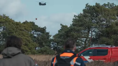 : Een helikopter met Bambi Bucket vliegt tussen de bomen. In de voorgrond lopen een militair en een brandweerman. Rechts staat een rode terreinwagen van de brandweer.