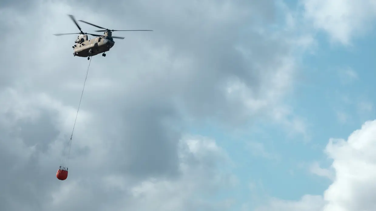 Helikopter met Bambi Bucket komt van links aanvliegen tegen een bewolkte lucht waar de zon op schijnt.