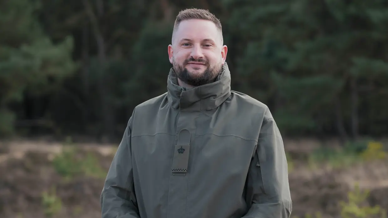 Een militair poseert met handen over elkaar op de hei.