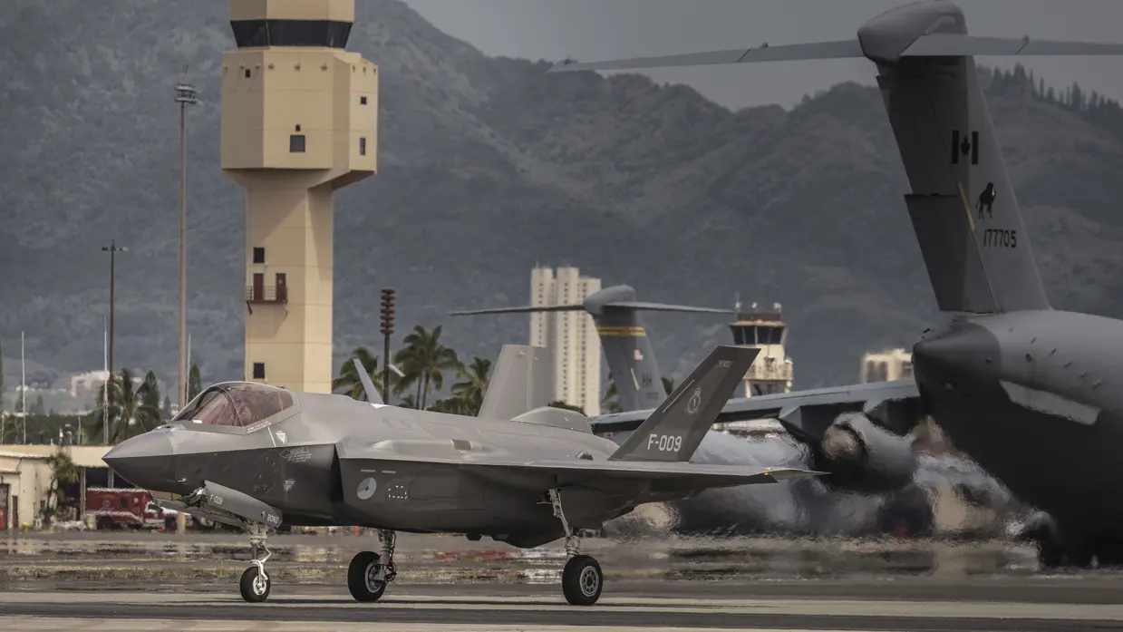 Een F-35 taxiet op Hawaii voor de verkeerstoren langs. In de achtergrond staan C-17 transportvliegtuigen van de VS en uit Canada