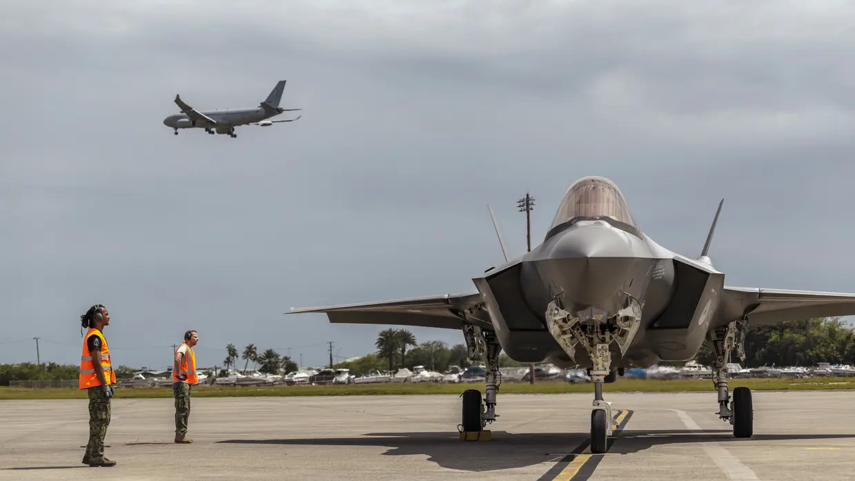 Een F-35 wordt door grondpersoneel met oranje hesjes naar zijn parkeerplaats geleid terwijl daarboven een A330 in de landing is te zien.