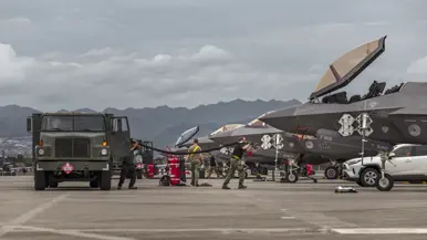 Luchtmachtpersoneel sjouwt brandstofslangen uit een Amerikaanse tankwagen naar een F-35. In de achtergrond zijn bergen te zien.