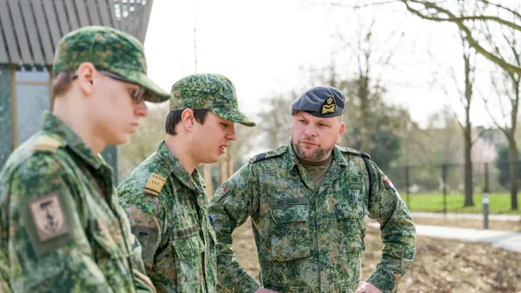 Sergeant-majoor Tim Nelissen in gevechtspak met blauwe baret luistert met zijn handen in zijn zij naar twee jongens in militair gevechtstenue. Ze staan op een verharde weg met groenstroken eromheen.