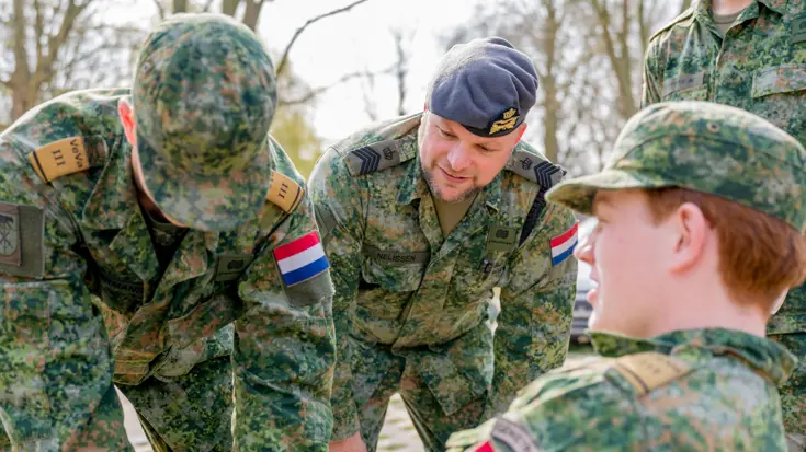 Een jonge militair zittend op een bankje, een andere met blauwe latexhandschoen aan buigt naar hem toe. In het midden van hen kijkt Tim toe.