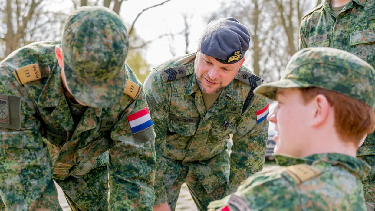 Een jonge militair zittend op een bankje, een andere met blauwe latexhandschoen aan buigt naar hem toe. In het midden van hen kijkt Tim toe.