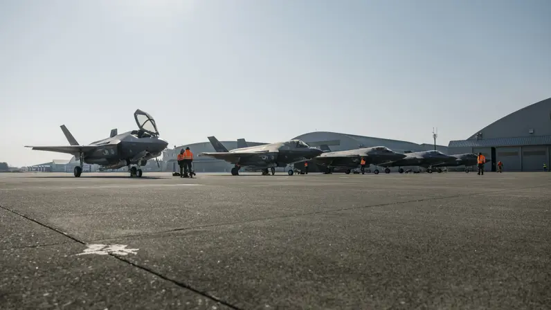 Alt-tekst: 5 F-35’s naast elkaar op een platform met in de achtergrond een aantal ronde hangers. Er staat personeel met oranje jassen tussen de F-35’s. 