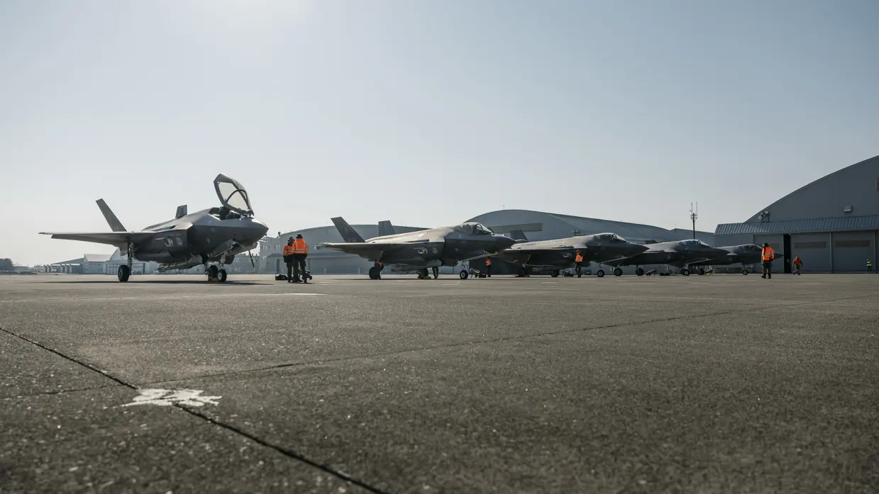 Alt-tekst: 5 F-35’s naast elkaar op een platform met in de achtergrond een aantal ronde hangers. Er staat personeel met oranje jassen tussen de F-35’s. 