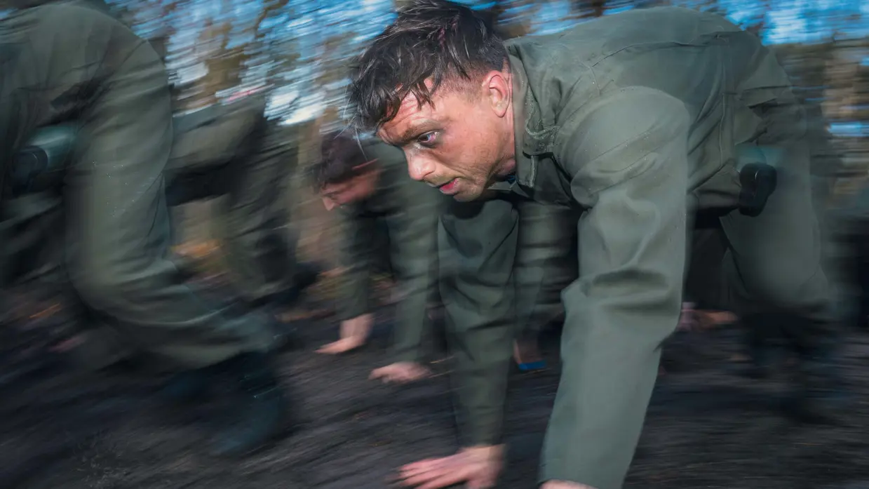 Een bezwete militair met camouflage onder het oog tijgert met collega’s door het bos.
