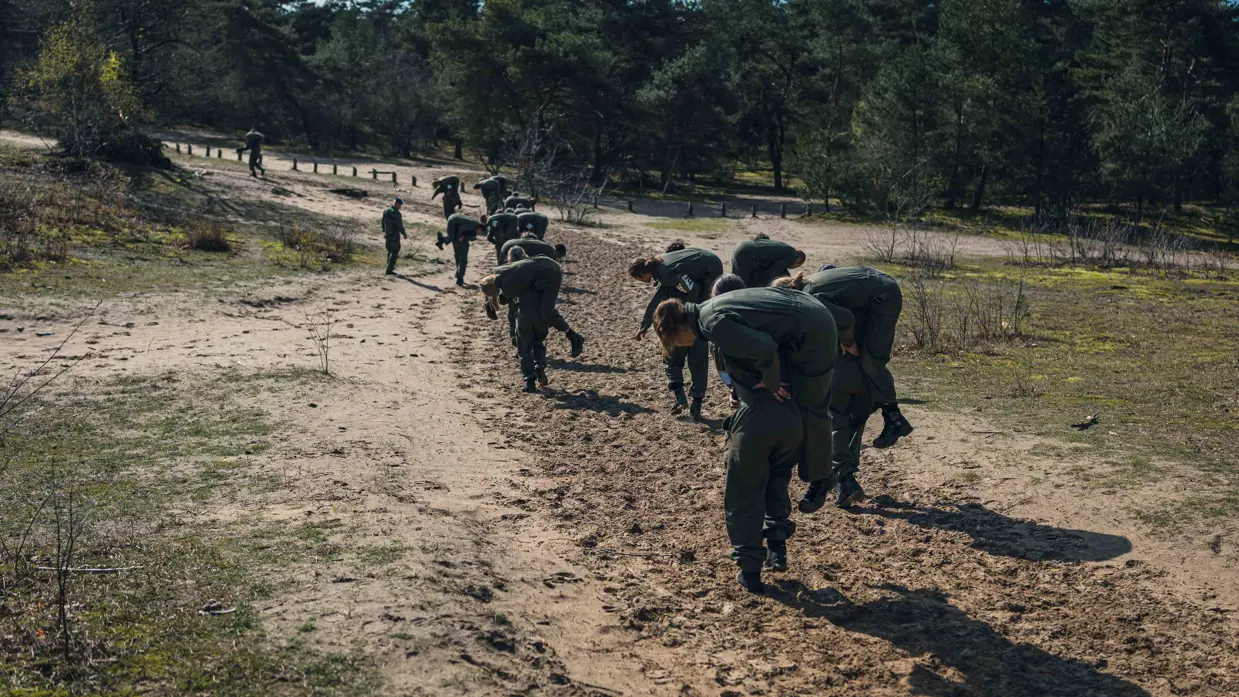 Meer dan 10 militairen rennen over de heide met een collega in de brandweergreep.