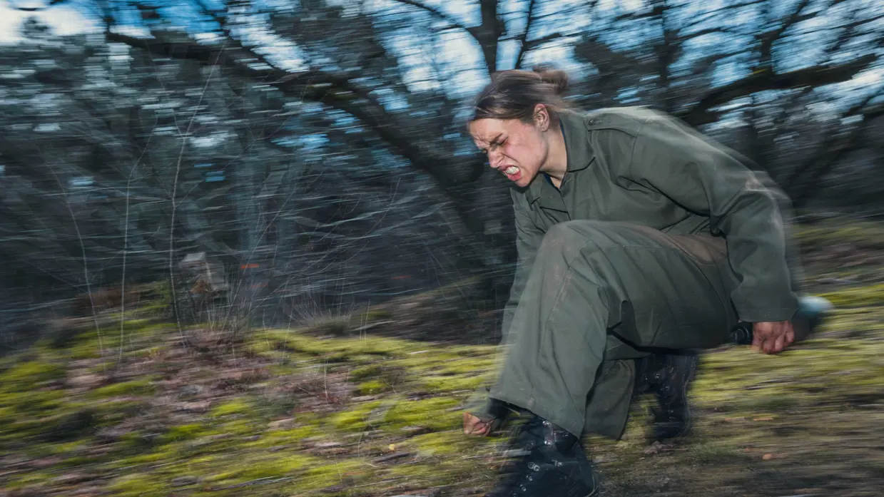 Een vrouwelijke cadet rent met een grimas op gezicht gehurkt door het bos.