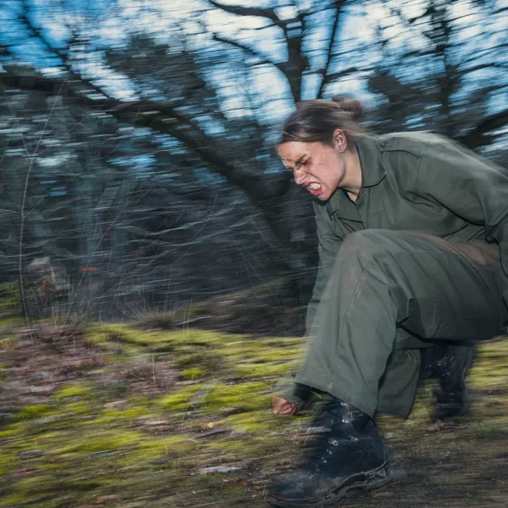 Een vrouwelijke cadet rent met een grimas op gezicht gehurkt door het bos.