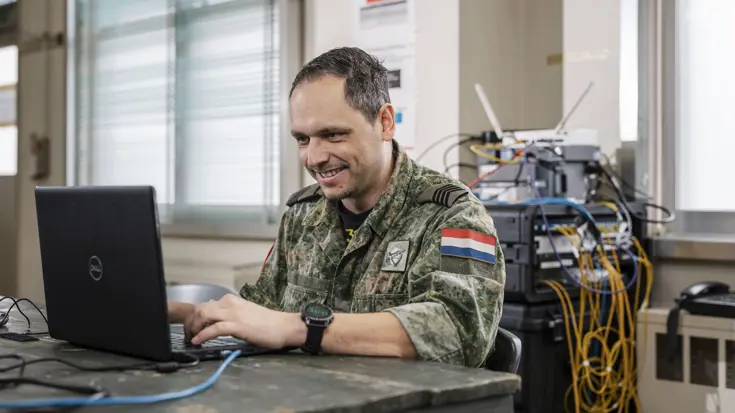 : Een militair in camouflagepak zit aan een tafel en werkt op een laptop. In de achtergrond is een wirwar van kabels te zien die uit diverse routers komen.