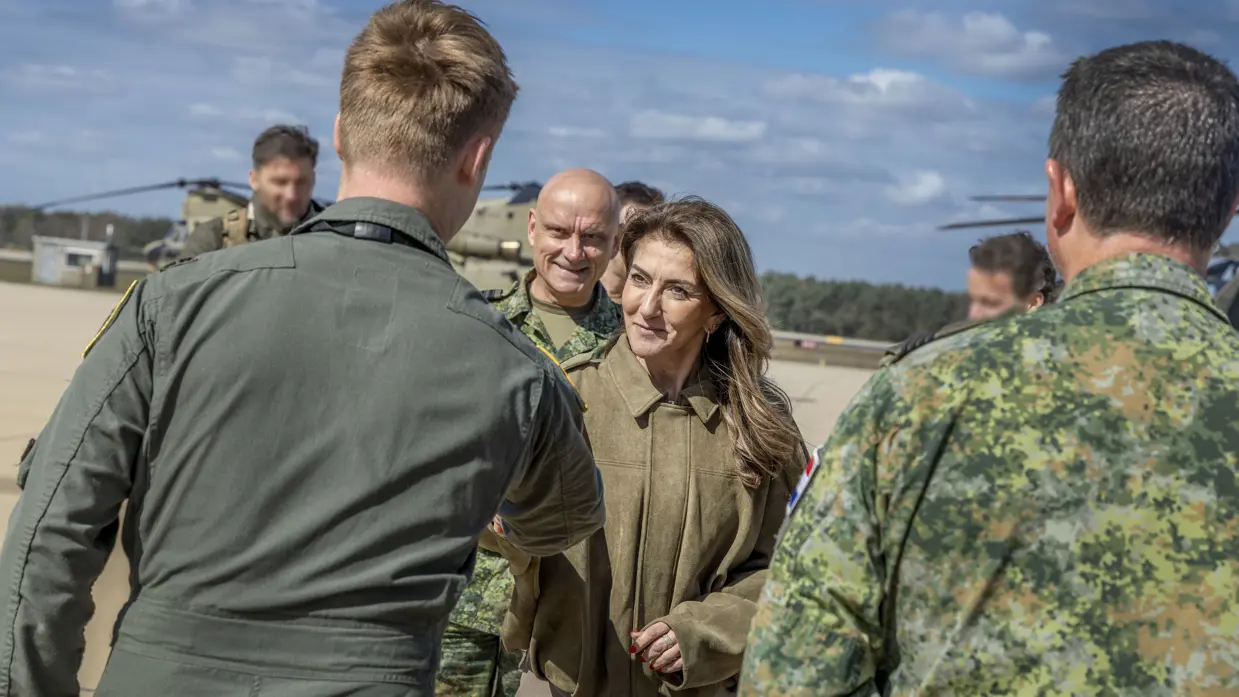 Buiten op een platform schudt de minister de hand van een militair in groene overall. Achter haar staat luitenant-generaal André Steur in gevechtstenue. Om hen heen staan meerdere militairen in gevechtstenue. Op de achtergrond staan Chinooks.