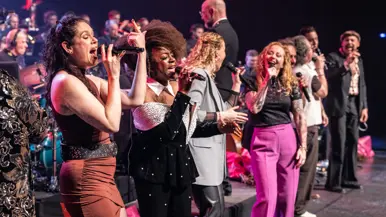 Diverse zangers en zangeressen voor op het podium met in de achtergrond het orkest met dirigent.