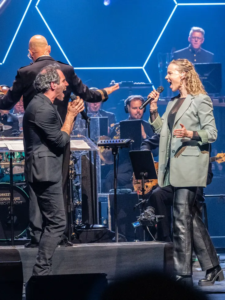 Diverse zangers en zangeressen voor op het podium met in de achtergrond het orkest met dirigent.