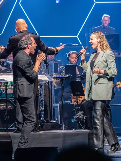 Diverse zangers en zangeressen voor op het podium met in de achtergrond het orkest met dirigent.