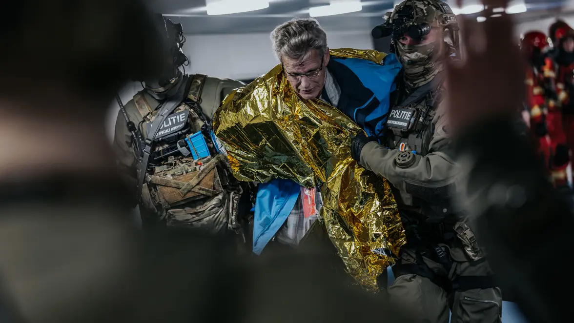 Tijdens een evacuatie heeft een gewonde man een gouden deken omgeslagen, terwijl twee operators hem ondersteunen. 