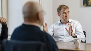 Twee mannen zitten aan een tafel in een lichte kamer. De man rechts, in wit overhemd, praat met handgebaren. Achter hem een raam. 