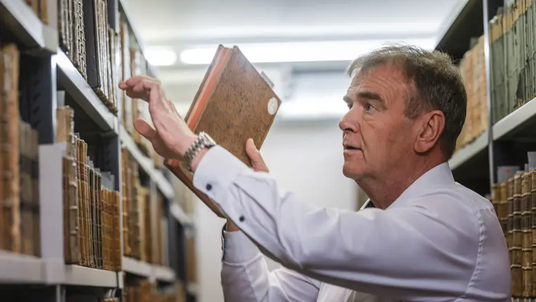 Een man in een wit overhemd pakt een oud, bruin boek van een plank vol oude boeken in een bibliotheek.