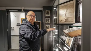 Een man met bril wijst naar een vitrine met oude documenten, foto's en een open boek in een museum.