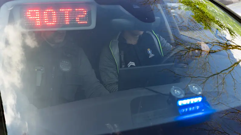 Twee Nederlandse marechaussees zitten in burger in een onopvallende auto, gezien door de voorruit.