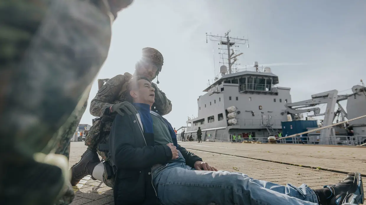 Een zogenaamde gewonde ligt op de grond en wordt geholpen door een militair. 