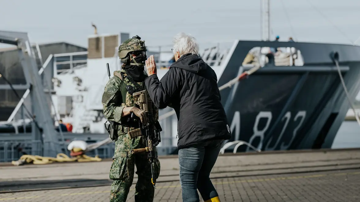 Een vrouw met een zwarte jas en blond haar loopt af op een militair in vlekkenpak. 
