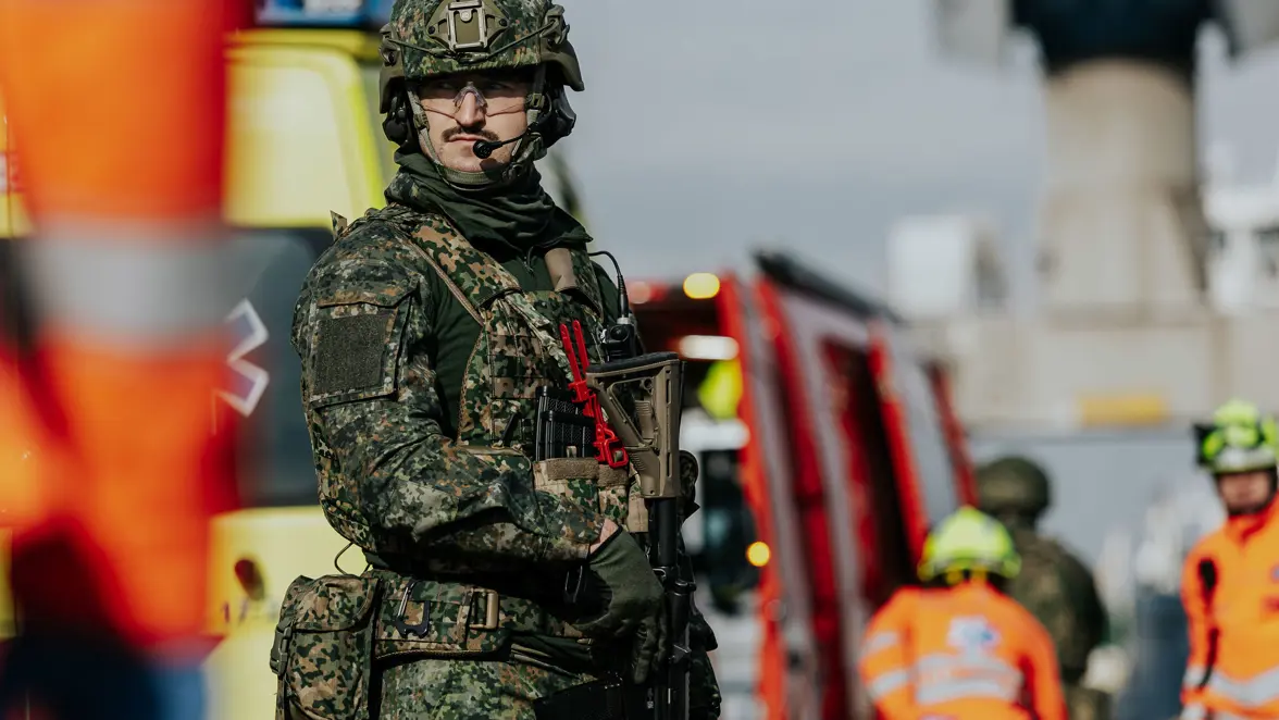 Een militair in groen vlekkenpak kijkt opzij. Achter hem staat personeel van de Brandweer. 