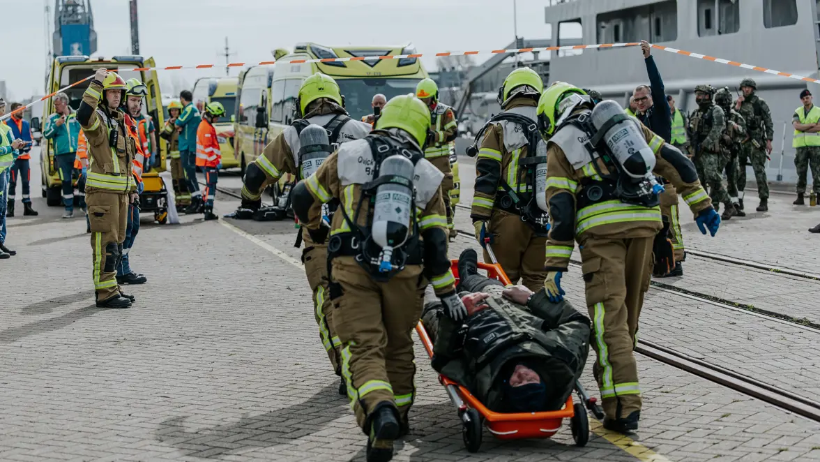 Een man in bompak wordt op een rijdende brancard afgevoerd door brandweerpersoneel. Daarachter is een menigte aan veiligheidspersoneel te zien, waaronder ambulancepersoneel en militairen. 