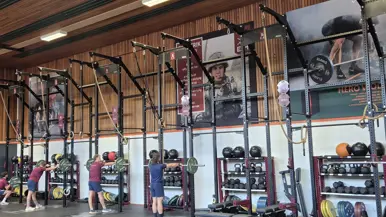 Een sportschool met krachttrainingsapparatuur. Er hangen gewichten, touwen en stangen. Vier mensen zijn aan het trainen, en de vloer is bedekt met rubberen matten.