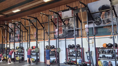 Een sportschool met krachttrainingsapparatuur. Er hangen gewichten, touwen en stangen. Vier mensen zijn aan het trainen, en de vloer is bedekt met rubberen matten.
