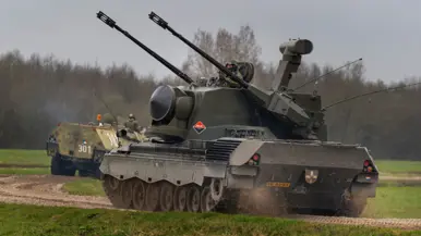 Een tank en gepantserde voertuigen rijden over een onverhard pad door een groen, open veld onder een bewolkte lucht. Rechts: Een zwaar militair voertuig met dubbele kanonnen en radar rijdt over een onverhard pad, gevolgd door andere legervoertuigen in een groen veld.