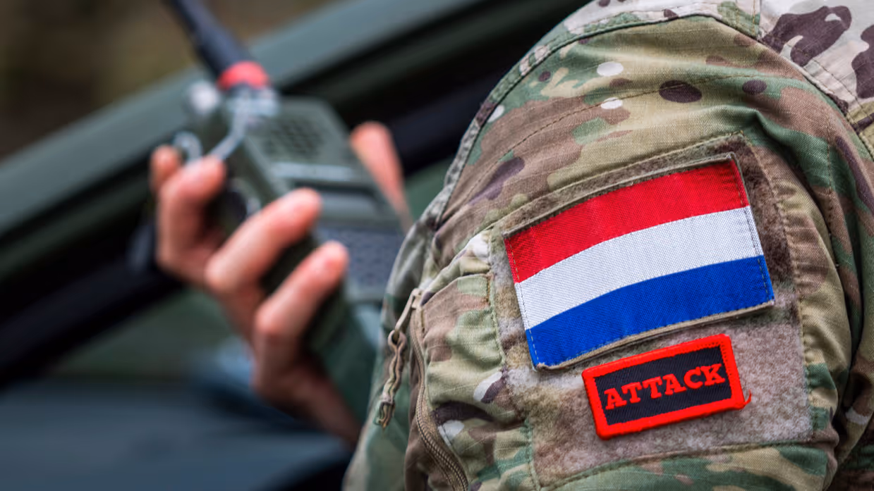 Een militair's linkerschouder, bedekt met patches, met in de achtergrond diens rechterhand die een walkietalkie vasthoudt.