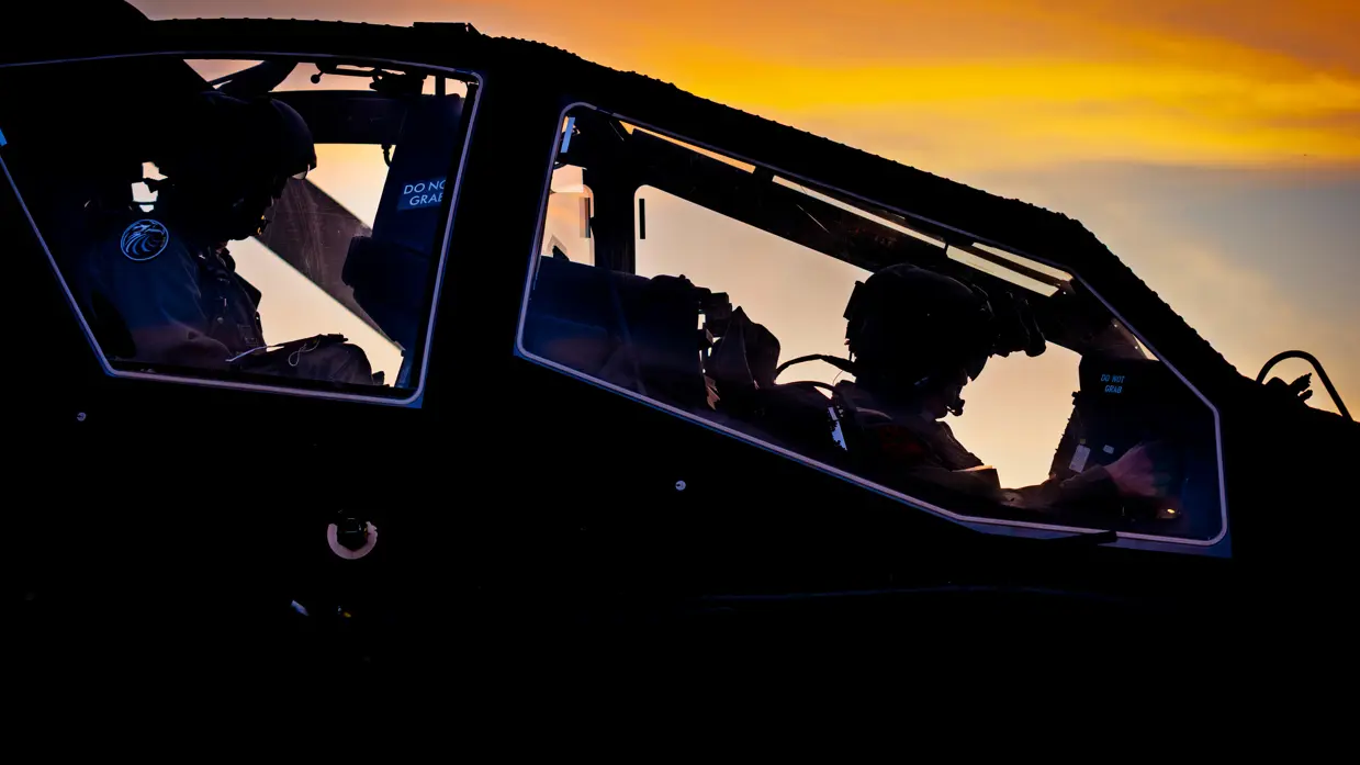 Een zijdelingse close-up van een Apache cockpit met twee piloten erin die van achter belicht worden door schemerend zonlicht.