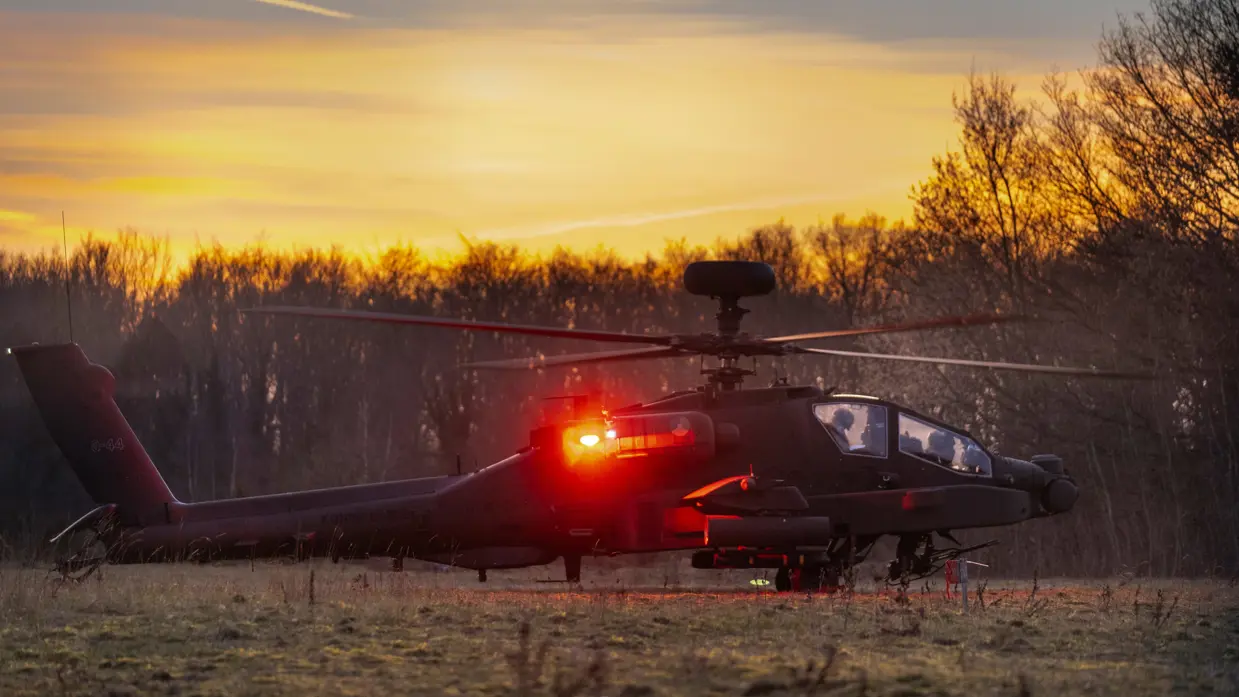 Een Apache-helikopter staat aan de grond met een fel rode reflectie op het toestel van het schemerlicht.