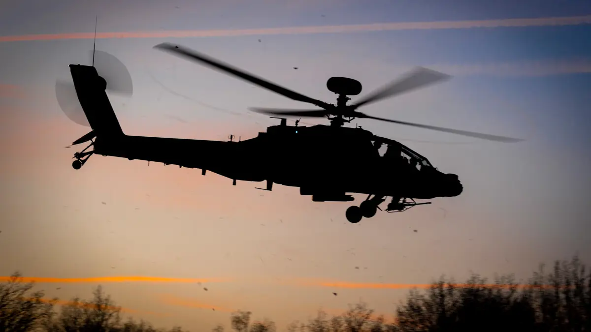 Een Apache-helikopter vliegt in de lucht met schemerend zonlicht in de achtergrond.