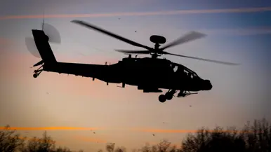Een militaire helikopter staat stil op een grasveld tijdens zonsondergang, met rode navigatielichten aan en bomen op de achtergrond. Rechts: Een silhouet van een militaire helikopter vliegt laag tijdens zonsondergang met draaiende rotorbladen tegen een gekleurde lucht met bomen op de achtergrond.
