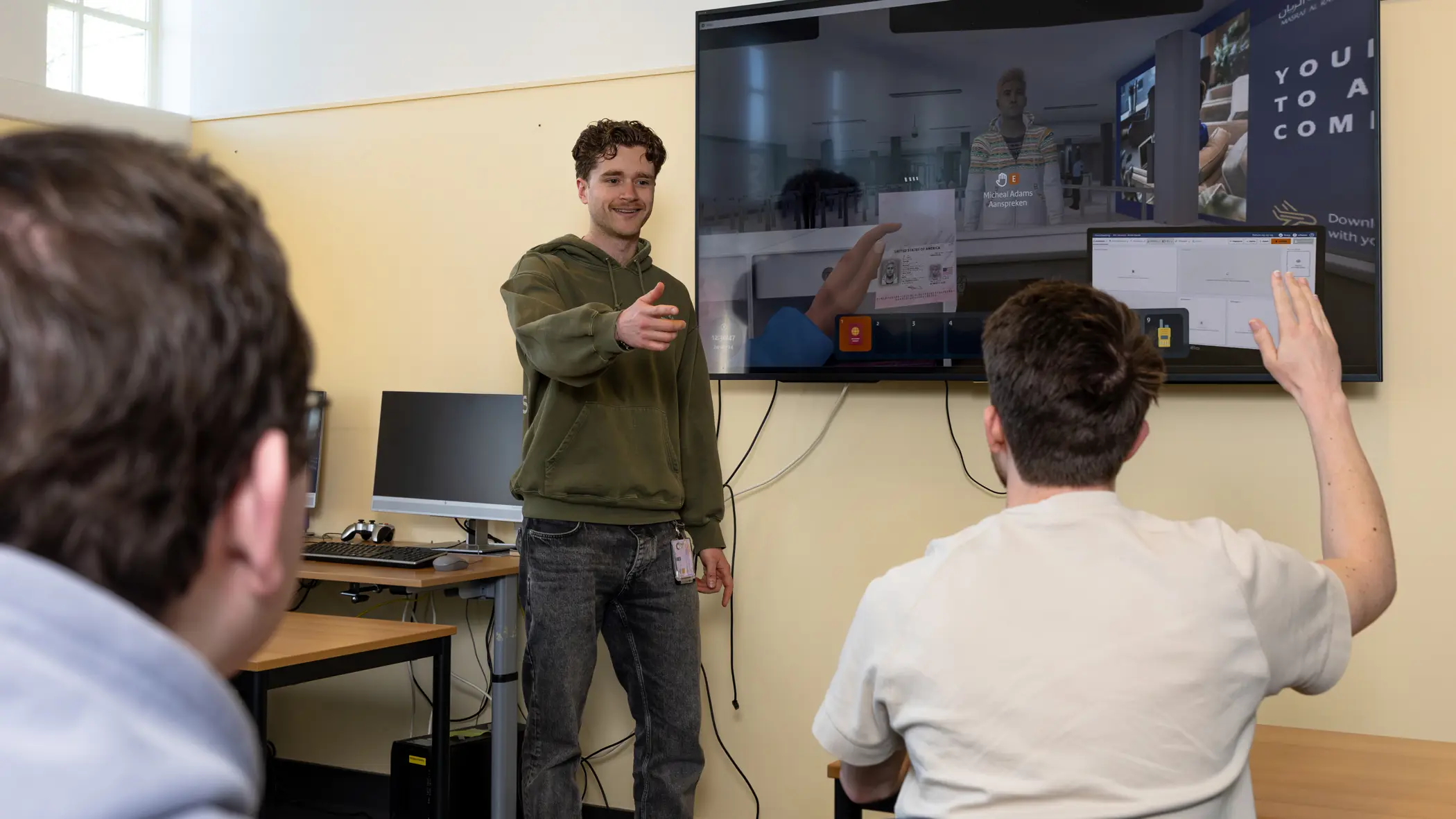 Een begeleider in een groene trui staat voor een digitaal schoolbord. Hij geeft de beurt aan een stagiair die zijn hand opsteekt.