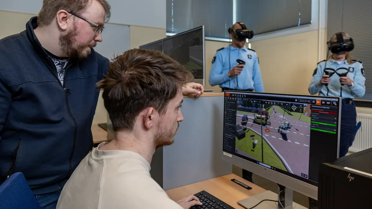 Twee mannen kijken naar een computerscherm waarop een animatie van een verkeerssituatie is te zien. Op de achtergrond hebben twee marechaussees een VR-bril op hun hoofd en controllers in hun handen.