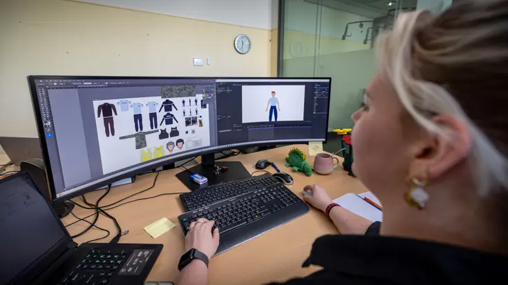 Een jonge vrouw kijkt naar een breed, holvormig computerscherm. Op het scherm is een animatie te zien van een marechausseepoppetje en verschillende soorten kleding. 