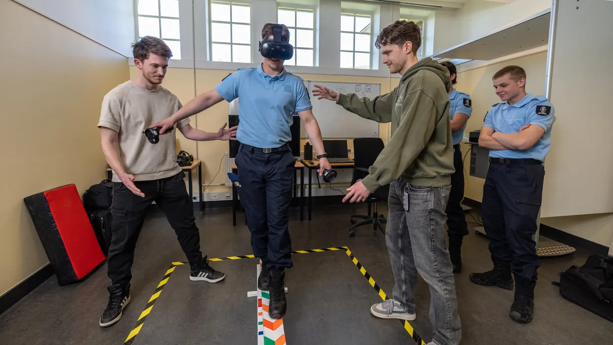 Een marechaussee in opleiding draagt een VR-bril en balanceert op een balk. Twee burgercollega’s begeleiden hem, terwijl op de achtergrond twee marechaussees toekijken.