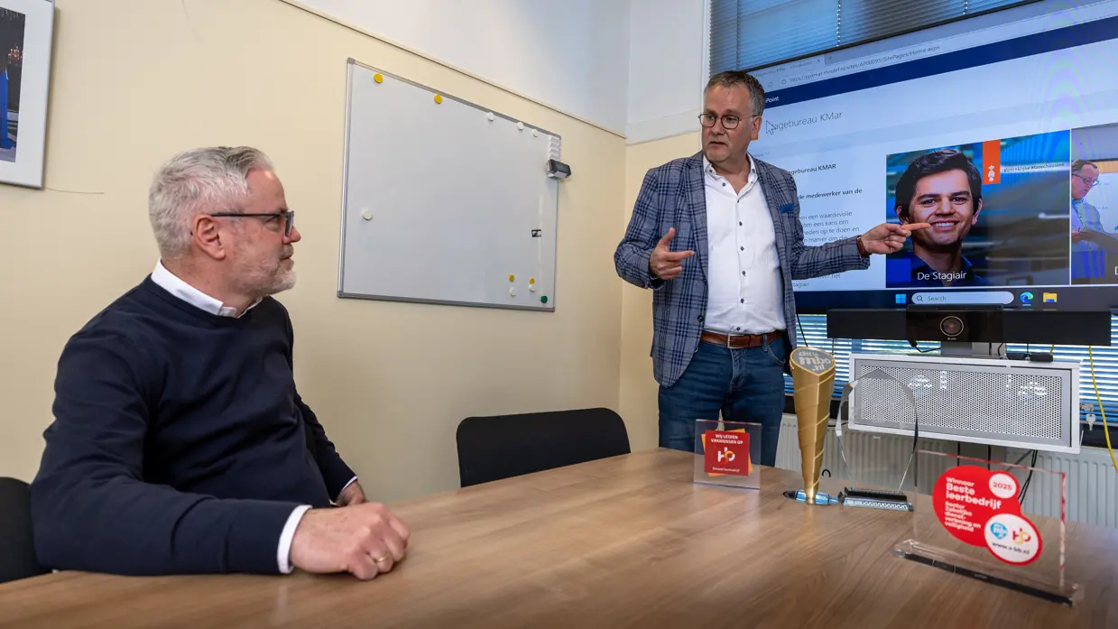 Een man staat bij een televisiescherm en wijst naar een foto van een stagiair. Links van hem zit een man te luisteren naar de uitleg. 